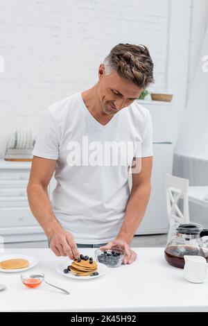 Mann, der frische Blaubeeren auf Pfannkuchen in der Nähe von Kaffee und Honig in der Küche legt, Stockbild Stockfoto