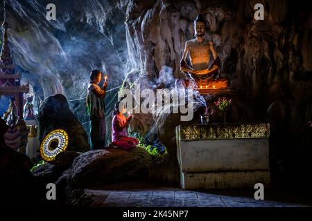Ein Höhlentempel mit Buddha-Statuen, brennenden Kerzen und zwei Mädchen, die knieten und beteten. Stockfoto