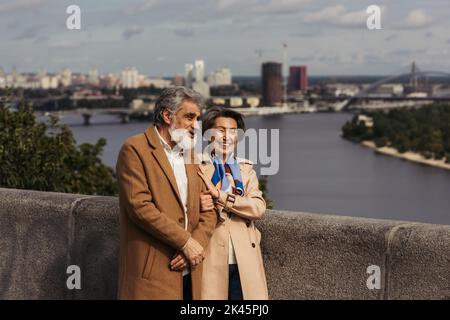 Glückliches Seniorenpaar in beigefarbenen Mänteln, lächelnd und auf der Brücke in der Nähe des Flusses, Stockbild Stockfoto