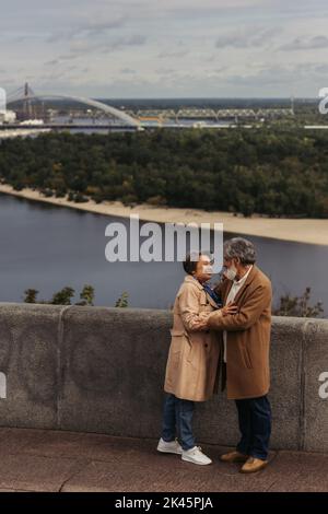 In voller Länge glückliches Seniorenpaar in beigen Herbstmänteln, umarmt auf Brücke in der Nähe des Flusses, Stockbild Stockfoto