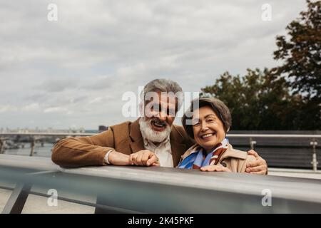 Bärtiger älterer Mann im Mantel, umarmt fröhliche Frau und steht in der Nähe der Brückenwache, Stockbild Stockfoto