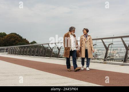 In voller Länge ein zufriedenes Seniorenpaar in beigen Mänteln, das die Hände hält und auf einer Brücke in der Nähe der Leitstange läuft, Stockbild Stockfoto