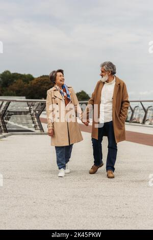 In voller Länge fröhliches Seniorenpaar in Mänteln, die die Hände halten und auf einer Brücke in der Nähe der Leitstange gehen, Stockbild Stockfoto