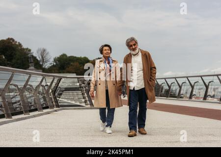 In voller Länge glückliches Seniorenpaar in Mänteln, die Hände halten und auf einer Brücke in der Nähe der Leitstange gehen, Stockbild Stockfoto