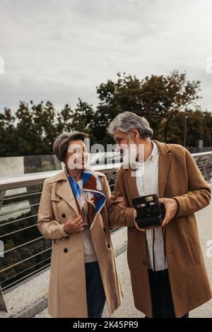 Glücklicher älterer Mann, der eine Vintage-Kamera in der Nähe einer lächelnden Frau auf der Brücke hält, Stockbild Stockfoto