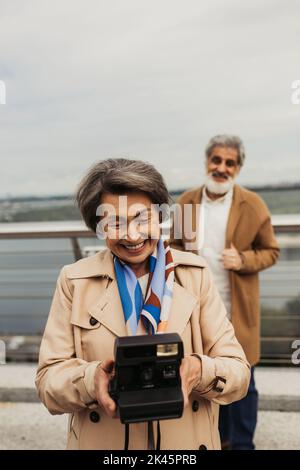 Glückliche ältere Frau hält Vintage-Kamera in der Nähe verschwommen Mann auf dem Hintergrund, Stock Bild Stockfoto