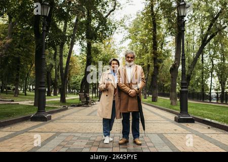 Lange ältere Frau hält Papierbecher, während sie mit stilvollem Ehemann im Mantel steht, Stockbild Stockfoto