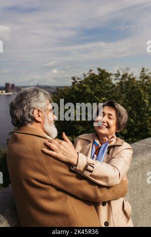 Glückliches und älteres Paar in beigefarbenen Mänteln, das sich auf der Brücke in der Nähe des Flusses umarmt, Stockbild Stockfoto