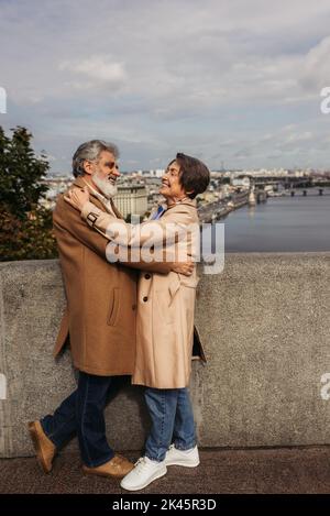 Glückliches und älteres Paar in beigefarbenen Mänteln, umarmt auf einer Brücke in der Nähe des Flusses, Stockbild Stockfoto