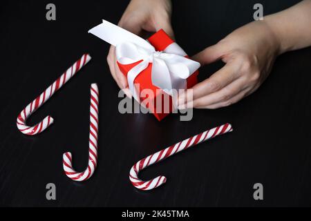 Rote Geschenkbox in weiblichen Händen auf dunklem Holztisch mit Zuckerstöcken. Konzept der Weihnachtsferien, romantische Überraschung Stockfoto