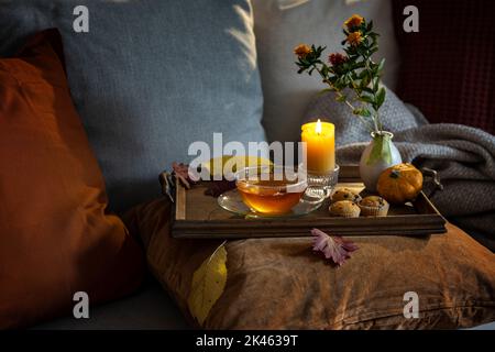 Gemütliche Teezeit im Herbst auf einem Tablett mit Kerze, Blumen und Herbstdekoration auf der Couch mit Kissen in warmen dunklen Farben serviert, Kopierraum, ausgewählt Stockfoto