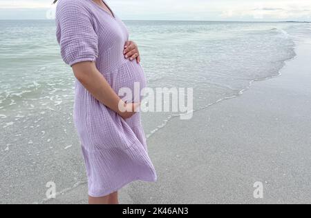 Aufnahme einer Schwangeren, die mit der Hand am Bauch am Strand steht - Schwangerschaft, Erwartung, Mutterschaft Konzept Stockfoto