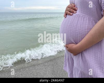 Aufnahme einer Schwangeren, die mit der Hand am Bauch am Strand steht - Schwangerschaft, Erwartung, Mutterschaft Konzept Stockfoto