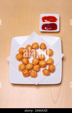 Gebratene Fischbällchen werden auf einen weißen Teller gelegt, auf einen Holztisch gestellt und mit Ketchup und Chilisauce serviert Stockfoto