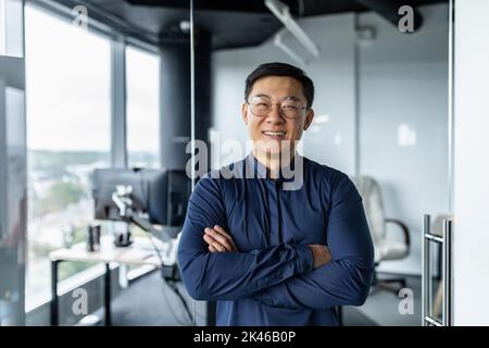 Porträt eines erfolgreichen asiatischen Geschäftsmannes mit gekreuzten Armen, eines Geschäftsmannes, der im Loft des Bürogebäudes arbeitet, die Kamera anschaut und einen lächelnden, zufriedenen Investor Finanzier zeigt. Stockfoto