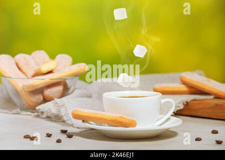 Weiße Tasse heißen aromatischen Kaffee mit Dampf und fallenden Zuckerwürfeln. Traditionelle italienische savoiardi-Kekse oder Marienkäfer auf einem Teller und in einem Stockfoto