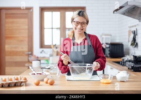 Schöne junge Frau mischt Teig, Blick auf die Kamera und lächelt beim Backen in der Küche zu Hause, Dekoration einen Kuchen aus Schokoladenkuchen, Kochen cl Stockfoto