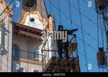 Kommunale Servicemitarbeiter in orangefarbenen Uniformen auf einer mobilen Luftplattform reparieren die festlichen elektrischen Girlanden des neuen Jahres. Stockfoto