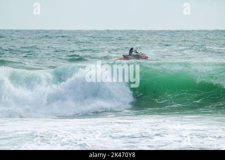 Eine große Welle azurblauen Wassers - im Hintergrund ein Mann auf einem Jetski. Er ist verantwortlich für die Sicherheit des Surfers beim Wettkampf (nicht erkennbarer pe Stockfoto