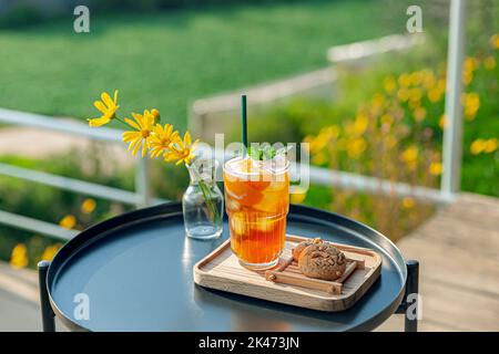 Frischer, hausgemachter Pfirsich-Eistee mit Minze, Schokoladenkekse auf dem Tisch im Freien. Sommerlicher, kalter Fruchtdrink am sonnigen Nachmittag mit gelben Blumen Stockfoto