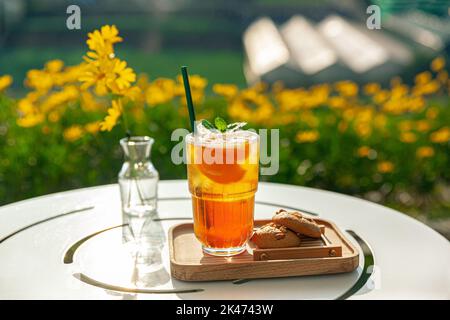 Frischer, hausgemachter Pfirsich-Eistee mit Minze, Schokoladenkekse auf dem Tisch im Freien. Sommerlicher, kalter Fruchtdrink am sonnigen Nachmittag mit gelben Blumen Stockfoto