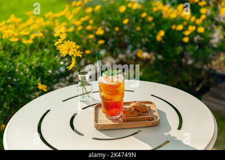 Frischer, hausgemachter Pfirsich-Eistee mit Minze, Schokoladenkekse auf dem Tisch im Freien. Sommerlicher, kalter Fruchtdrink am sonnigen Nachmittag mit gelben Blumen Stockfoto