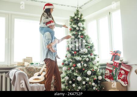 Familie schmücken einen Weihnachtsbaum. Junger Mann mit seiner Tochter auf seinen Schultern hilft ihr, den Weihnachtsbaum schmücken. Stockfoto