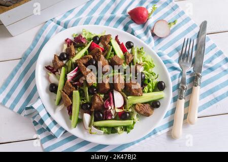 Gehacktes Rindfleisch, gekochte Stücke mit Salatmischung, grünen Bohnen, Gurken, Oliven und Radieschen. Auf einem weißen Teller. Auf einer gestreiften weiß-blauen Serviette. Gabel und kni Stockfoto