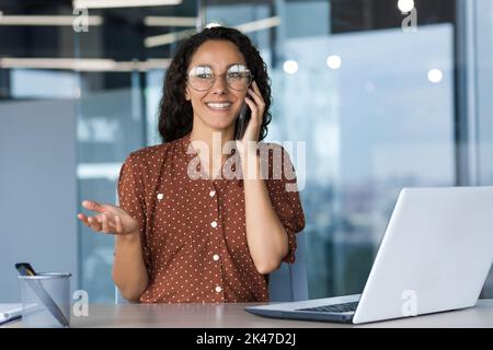 Junge, schöne hispanische Freiberuflerin, die eine Brille trägt und im Büro am Schreibtisch mit einem weißen Laptop sitzt und am Telefon spricht. Er lächelt, streicht mit den Händen. Stockfoto