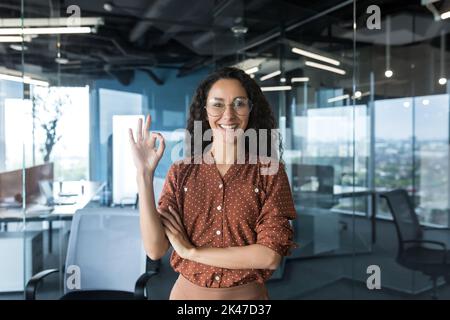 Porträt einer jungen schönen hispanischen Freiberuflerin, IT-Mitarbeiterin, SEO in Brille steht im Büro, schaut auf die Kamera, lächelt, zeigt ok mit ihrer Hand. Stockfoto