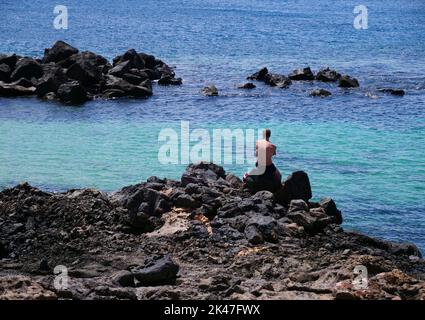 Der Mann sitzt mit dem Rücken auf einem Felsen und blickt entspannt auf das Meer Stockfoto