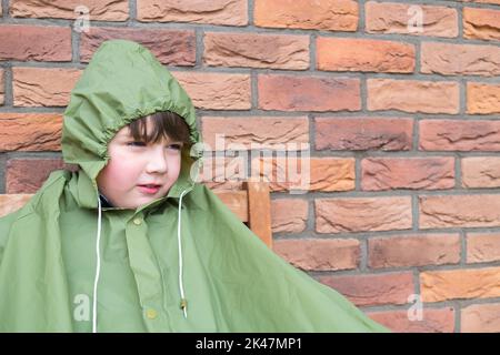 Kind, Junge in der grünen Regenjacke sitzt und wartet auf Stopp regnen. Regenzeit im Herbst. Backsteinmauer Hintergrund. Regnerisches Tageswetter. Stockfoto