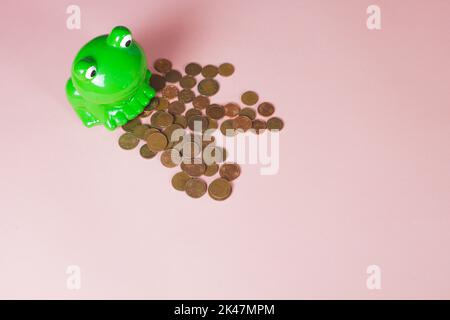 Keramik-Geldkasten, Form und Farbe des grünen Frosches. Euro-Münzen, die von der Kasse auf dem rosa Hintergrund mit Platz für Text ausgegossen wurden. Sparschwein. Stockfoto