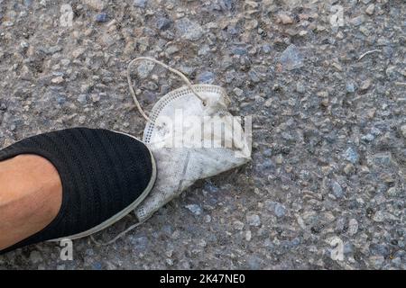 Nahaufnahme eines weißen schmutzigen gebrauchten Atemschutzgeräts mit Fuß in schwarzem Schuh, der auf grauem Bürgersteig steht. Konzept des Endes der Covid-19-Pandemie, Freiheitsbeschränkung. Stockfoto