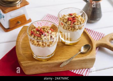 Müsli mit Mangosauce und Goji-Beeren. Auf einem weißen Holzbrett, einem Eisenlöffel, einer Kaffeekocher, einer türkischen Kaffeekocher, einem weißen Holztisch und einer Serviette Stockfoto