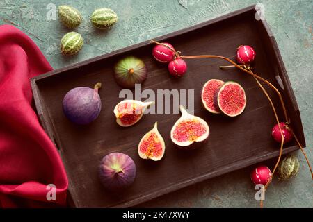 Herbstzeit Hintergrund mit frischen halbierten Feigenfrüchten auf schwarzem Holztablett. Himbeerroter Seidenschal und Dekofrüchte auf türkis, mintgrün strukturiert Stockfoto