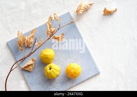 Buntes Herbstmuster mit gelben Quitten-Früchten auf blauer Keramikfliese. Saisonales Stillleben im Herbst auf einer weißen Textiltischplatte aus Baumwolle. Trocken Stockfoto