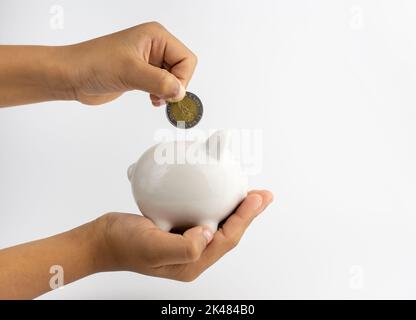 Ein Sparschwein in der Hand und setzt Münzen in ein Sparschwein mit Kopierraum auf weißem Hintergrund. Stockfoto