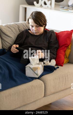 Ein unwohl vorjugendlicher Junge, der auf einer Couch liegt und auf seine Tablette schaut, seine Wangen sind von einem Fieber rot und er hat eine Gewebebox neben sich Stockfoto