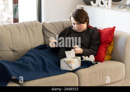 Ein unwohl vorjugendlicher Junge, der auf einer Couch liegt und auf seine Tablette schaut, seine Wangen sind von einem Fieber rot und er hat eine Gewebebox neben sich Stockfoto