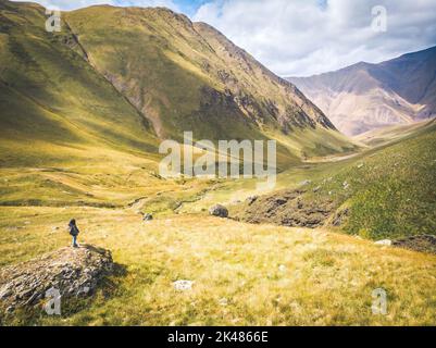 Die Backpacker-Frau steht auf dem Felsen und genießt einen atemberaubenden Blick auf die Berge im Juta-Tal. Erkundung des Nationalparks Kazbegi. Im Freien im kaukasus frisch Stockfoto