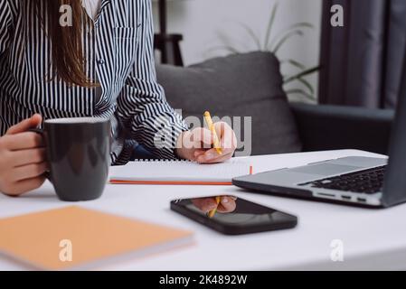 Nahaufnahme der weiblichen Hände, die Tasse Kaffee und Stift halten und Notizen in Tagebuch machen, sitzen Sie mit einem Laptop am Schreibtisch. Geschäftsfrau, die bei der Arbeit einen Geschäftsbrief schreibt. Kostenlos Stockfoto