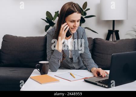 Nette lächelnde junge Frau im Alter von 20s Jahren, die am Schreibtisch neben einer heißen Kaffeetasse auf einer bequemen Couch telefoniert und am Laptop arbeitet Stockfoto