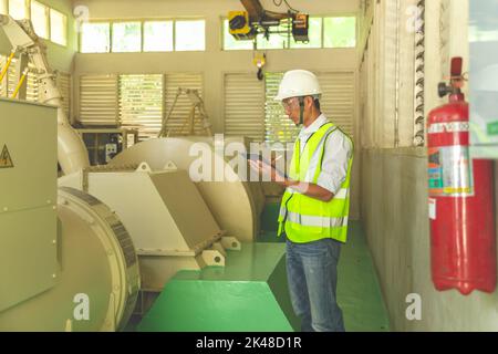 Männlicher Superyacht-Ingenieur, der im Maschinenraum arbeitet und den Generator mit einem Checkliste-Ordner und einem Stift in der Hand inspiziert. Stockfoto