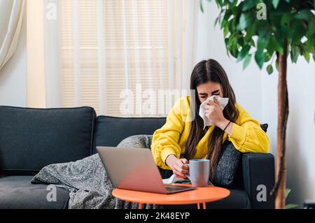 Kranke junge Frau in gelbem Sweatshirt, trinkt warmen Tee, bläst ihre laufende Nase, mit Laptop. Stockfoto