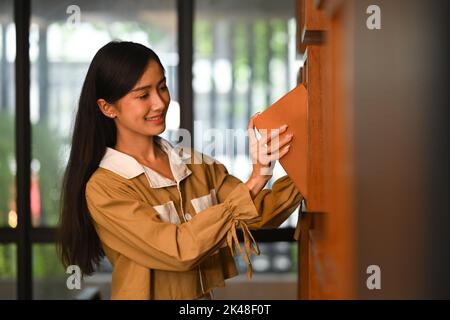 Hübsche asiatische Studentin, die ein Buch aufnimmt und Bücher aus dem Regal in der Universitätsbibliothek wählt Stockfoto
