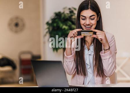 Enthusiastische junge Brünette, die ihre Debitkarte auscheckte, im Büro saß und ihren Laptop benutzte. Stockfoto