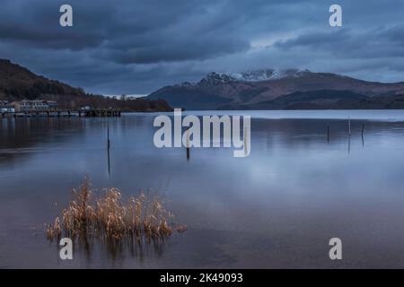 Atemberaubendes Landschaftsbild von Loch Lomond und schneebedeckter Bergkette in der Ferne vom kleinen Dorf Luss aus gesehen Stockfoto