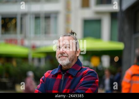 Ein Mann mittleren Alters mit einem chaotischen Brötchen in einer rot-blauen Jacke im Stil eines Holzfällers steht mit gekreuzten Armen in einer Einkaufsstraße Stockfoto