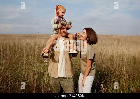 Glückliche Familie, die im Herbst oder Sommer im Freien Spaß hat. Tochter sitzt auf Vater, Mutter lacht. Lifestyle, Natur beige Feld Stockfoto